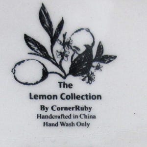 May include: A white ceramic surface featuring a black ink illustration of a lemon and leaves. The text reads: The Lemon Collection, By CornerRuby, Handcrafted in China, Hand Wash Only. The design is centered and printed in a clear, dark font.