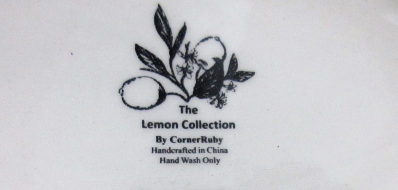 May include: A white ceramic surface featuring a black ink illustration of a lemon and leaves. The text reads: The Lemon Collection, By CornerRuby, Handcrafted in China, Hand Wash Only. The design is centered and printed in a clear, dark font.