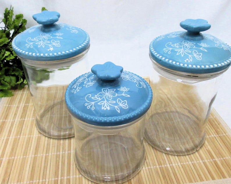 May include: Three glass canisters with blue and white floral patterned ceramic lids. The canisters are clear glass and the lids have a white floral design on a light blue background.