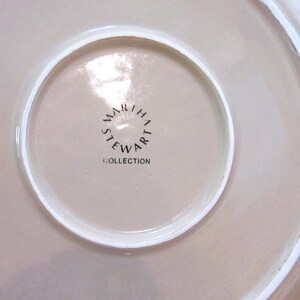 以下が含まれることがあります： ベージュ色の皿のクローズアップ。白い縁取りがあります。皿の中央には、黒字で「MARTHA STEWART COLLECTION」と書かれており、円形に配置されています。