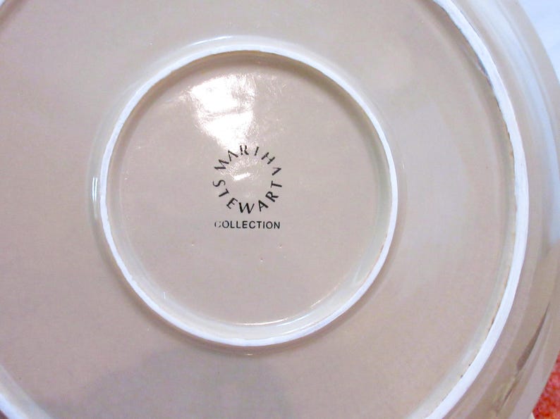 以下が含まれることがあります： ベージュ色の皿のクローズアップ。白い縁取りがあります。皿の中央には、黒字で「MARTHA STEWART COLLECTION」と書かれており、円形に配置されています。
