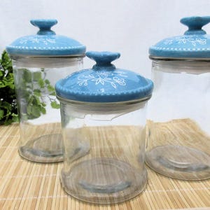 Puede incluir: Tres botes de vidrio con tapas de cerámica azul y blanca. Las tapas tienen un diseño floral.