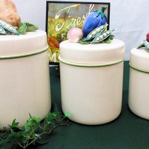 Könnte beinhalten: Drei cremefarbene Keramikbehälter mit grünen Rändern, die jeweils mit Keramikgemüse belegt sind. Der größte hat eine Kartoffel, der mittlere eine Aubergine und der kleinste einen roten Kürbis. Im Hintergrund ein gerahmtes Bild mit dem Wort "Fresh".