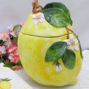 May include: A yellow ceramic lemon-shaped canister with a removable lid. The surface features a textured rind finish, decorated with raised green leaves, brown stems, and white flowers with pink centers. The canister has a rounded, bulbous shape.