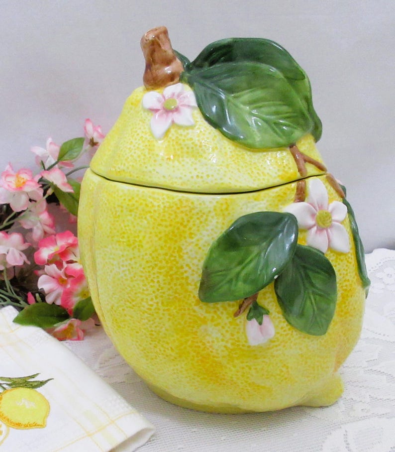May include: A yellow ceramic lemon-shaped canister with a removable lid. The surface features a textured rind finish, decorated with raised green leaves, brown stems, and white flowers with pink centers. The canister has a rounded, bulbous shape.