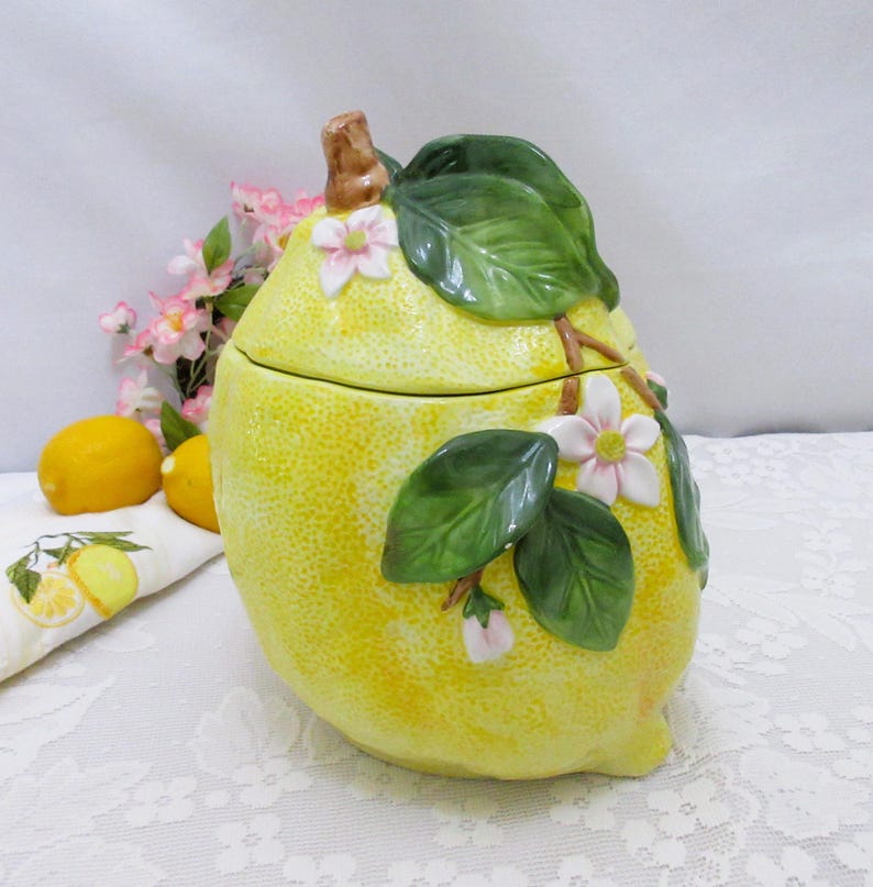 May include: A ceramic lemon-shaped canister with a textured yellow surface, featuring a removable lid with a brown stem, glossy green leaves, and small white and pink blossoms. The container has a rounded base and a realistic citrus fruit design.