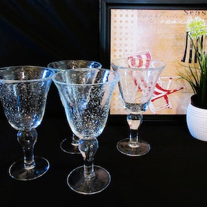 May include: Four clear, stemmed wine glasses with a bubble-like texture. The glasses have a flared bowl shape and a round base. A framed print with beach chairs and the words "coast relax beach" is in the background.