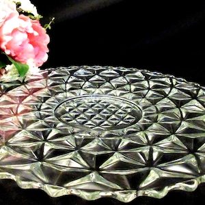 Może przedstawiać: Przezroczysty szklany talerz do serwowania z geometrycznym wzorem w kształcie diamentu. Talerz ma falisty brzeg i podniesiony środek. Różowe i białe kwiaty w tle.