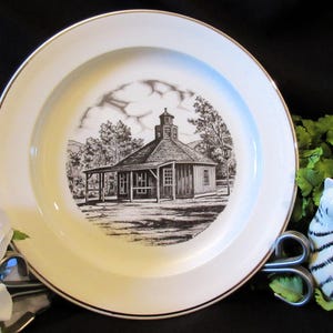 Puede incluir: Un plato de cerámica blanco con un borde dorado presenta una ilustración en blanco y negro de un edificio con un campanario. El edificio está rodeado de árboles y una valla. El plato es un objeto de colección vintage.