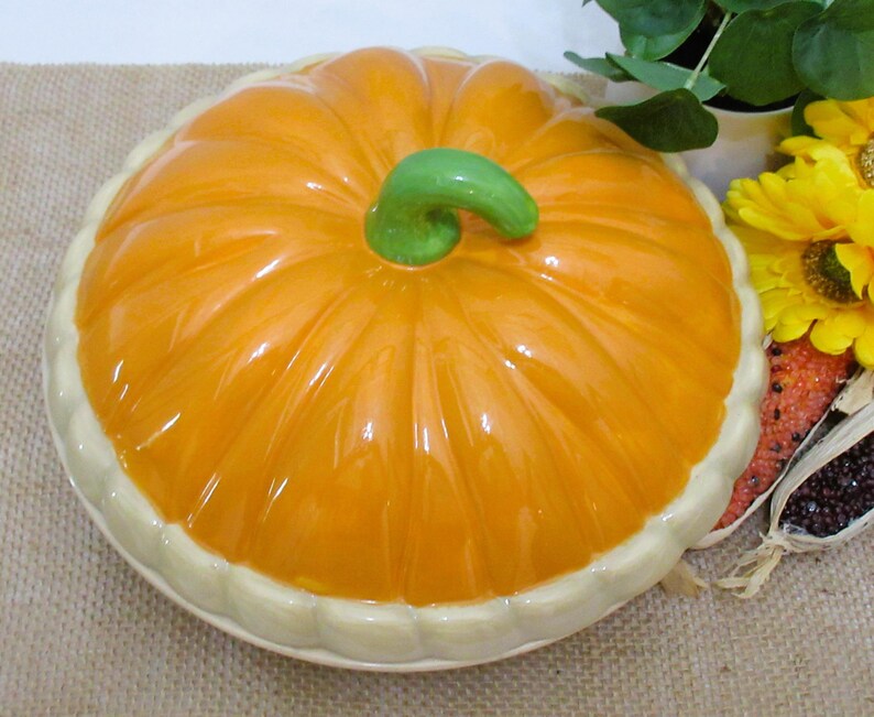 以下が含まれることがあります： 光沢のあるオレンジ色の仕上げと緑色の茎が付いた、カボチャ型のセラミック製サービングディッシュ。 ディッシュはクリーム色のトリムが付いたスカラップエッジです。 秋の季節の装飾品です。