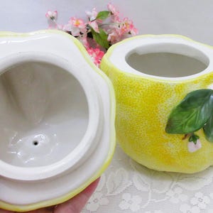 May include: A ceramic lemon-shaped jar with a removable lid. The exterior features a textured yellow surface with three-dimensional green leaves and small white flowers with yellow centers. The interior of the jar and the underside of the lid are white.