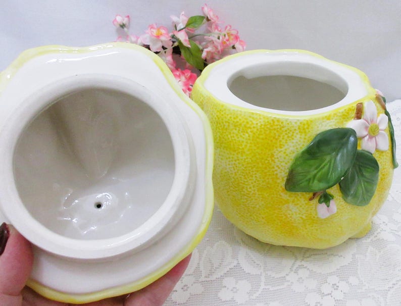 May include: A ceramic lemon-shaped jar with a removable lid. The exterior features a textured yellow surface with three-dimensional green leaves and small white flowers with yellow centers. The interior of the jar and the underside of the lid are white.