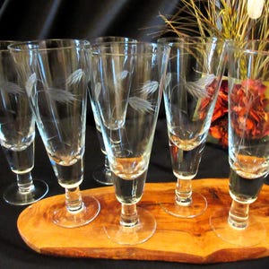 May include: Six clear champagne flutes with etched feather designs are displayed on a wooden serving tray. The glasses have a slender, tapered shape and a round base. The tray is oval-shaped and made of light-colored wood.