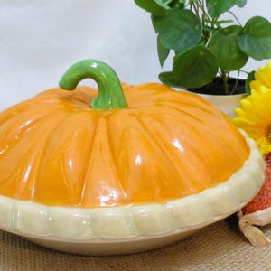 以下が含まれることがあります： 緑色の茎とクリーム色のベースが付いた、オレンジ色のセラミックカボチャ型のサービングディッシュ。皿はジュートの表面にあり、近くに黄色い花と装飾用のトウモロコシがあります。秋の装飾品。