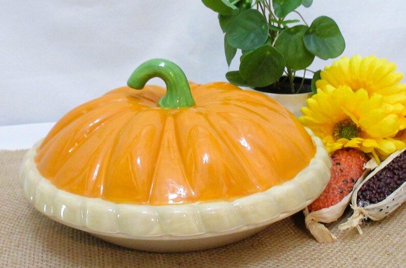 以下が含まれることがあります： 緑色の茎とクリーム色のベースが付いた、オレンジ色のセラミックカボチャ型のサービングディッシュ。皿はジュートの表面にあり、近くに黄色い花と装飾用のトウモロコシがあります。秋の装飾品。
