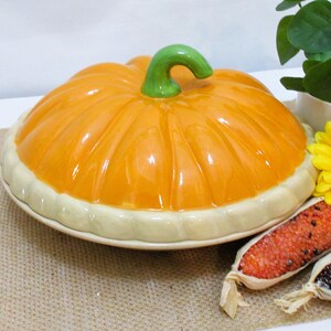 以下が含まれることがあります： 明るいオレンジ色のトップと緑色の茎が付いた、カボチャ型のセラミック製サービングディッシュ。 ディッシュはクリーム色の縁があり、ジュートの表面に置かれています。 黄色の花と装飾用のトウモロコシも存在します。