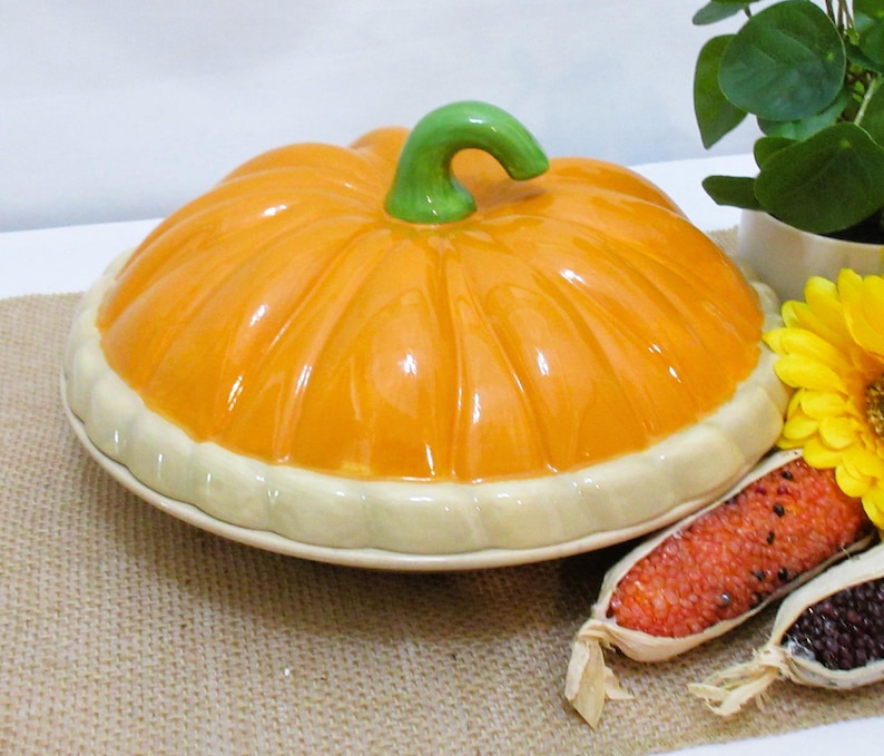 以下が含まれることがあります： 明るいオレンジ色のトップと緑色の茎が付いた、カボチャ型のセラミック製サービングディッシュ。 ディッシュはクリーム色の縁があり、ジュートの表面に置かれています。 黄色の花と装飾用のトウモロコシも存在します。