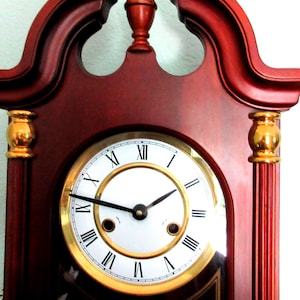 May include: A wooden grandfather clock with a white clock face and gold trim. The clock face features black Roman numerals and black hands. The clock's frame is a dark reddish-brown color with gold accents.