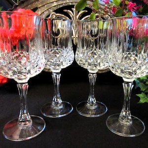 Puede incluir: Cuatro copas de vino de cristal transparente con un patrón de corte de diamante en el cuenco y un tallo delgado. Las copas están dispuestas en fila, mostrando su elegante diseño. El fondo incluye flores y una pieza metálica decorativa.