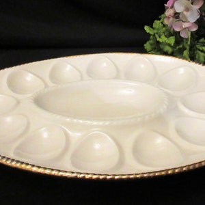 May include: A white ceramic egg plate with a gold rim and 12 individual egg-shaped depressions. The plate has a central oval-shaped depression for dipping sauce.