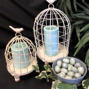 Op de afbeelding: Twee witte metalen vogelkooien met blauwe kaarsen erin. De vogelkooien hebben kleine vogelbeeldjes bovenop. Een kom met blauwe gevlekte eieren staat voor de vogelkooien.