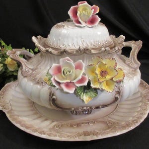 Puede incluir: Una sopera de cerámica blanca con tapa y plato a juego. La sopera está adornada con detalles florales rosas y amarillos. Las asas y los bordes tienen un ribete marrón. La sopera se encuentra sobre un plato a juego.