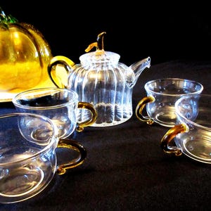 Puede incluir: Un juego de té de cristal transparente con una pieza decorativa en forma de calabaza. El juego incluye una tetera y cuatro tazas de té con asas doradas. La calabaza es dorada y está sobre un plato blanco.