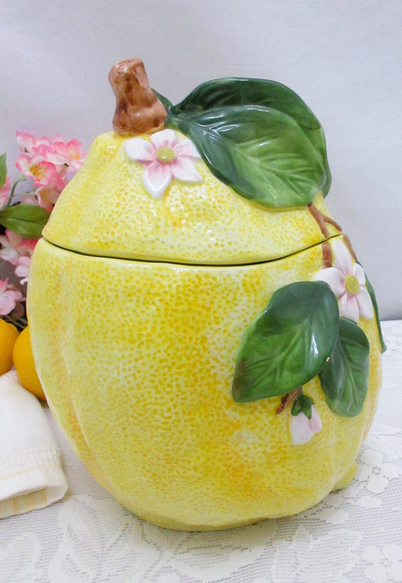 May include: A ceramic cookie jar shaped like a large yellow lemon with a textured rind. The lid features a brown stem and green leaves with small white and pink blossoms. The body of the jar also displays green leaves and white flowers.