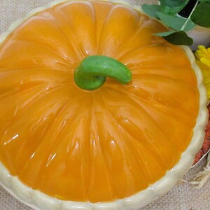 以下が含まれることがあります： 光沢のあるオレンジ色の仕上げと緑色の茎が付いた、カボチャ型のセラミック製サービングディッシュ。白い縁があり、ヒマワリやトウモロコシを使った秋をテーマにした装飾の一部です。