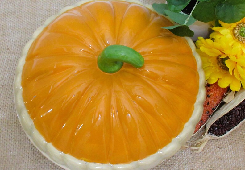 以下が含まれることがあります： 光沢のあるオレンジ色の仕上げと緑色の茎が付いた、カボチャ型のセラミック製サービングディッシュ。白い縁があり、ヒマワリやトウモロコシを使った秋をテーマにした装飾の一部です。