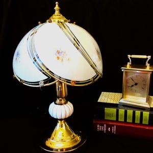 Pode incluir: Uma luminária de mesa vintage com acabamento branco e dourado. A luminária apresenta uma cúpula com detalhes florais e acabamento dourado. Um pequeno relógio dourado está em uma pilha de livros.