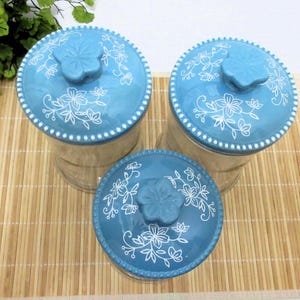May include: Three blue glass canisters with white floral designs and blue lids. The canisters are on a bamboo mat.