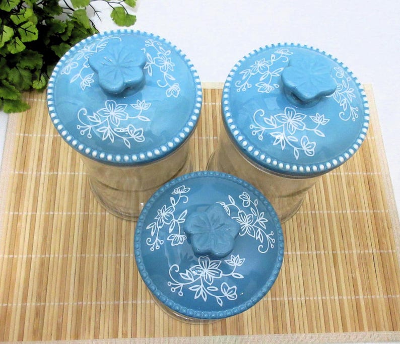 May include: Three blue glass canisters with white floral designs and blue lids. The canisters are on a bamboo mat.