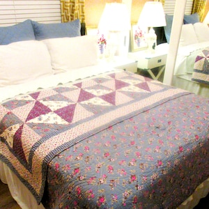 May include: A bed featuring a quilt with a blue floral pattern and a section of purple and white geometric patchwork. The bed includes two white pillows and two blue pillows, with a white bed skirt visible at the base of the mattress frame.
