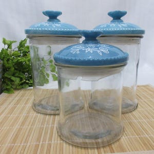 May include: Set of three glass canisters with blue and white floral patterned lids. The canisters are clear glass and the lids are ceramic.