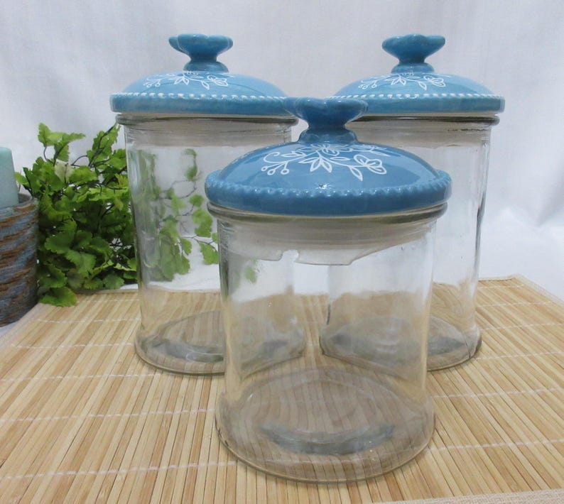 May include: Set of three glass canisters with blue and white floral patterned lids. The canisters are clear glass and the lids are ceramic.