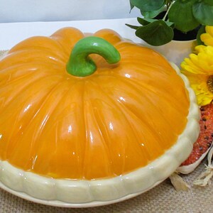 以下が含まれることがあります： 緑色の茎とクリーム色の縁が付いた、カボチャ型のセラミック製サービングディッシュ。オレンジ色の皿は、黄色い花と装飾用のトウモロコシの隣にあります。秋や感謝祭に最適です。