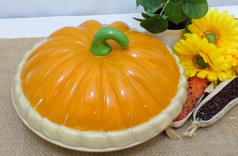 以下が含まれることがあります： 緑色の茎とクリーム色の縁が付いた、カボチャ型のセラミック製サービングディッシュ。オレンジ色の皿は、黄色い花と装飾用のトウモロコシの隣にあります。秋や感謝祭に最適です。