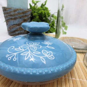 May include: A blue ceramic lid with a white floral design. The lid has a raised knob in the centre and a beaded edge.