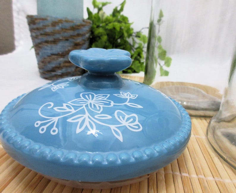 May include: A blue ceramic lid with a white floral design. The lid has a raised knob in the centre and a beaded edge.