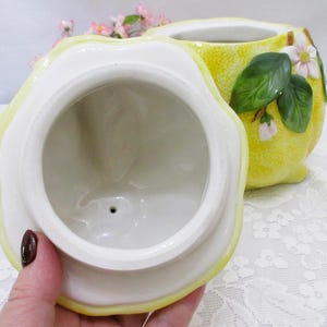 May include: A ceramic lemon-shaped jar with a removable lid. The exterior features a textured yellow rind, green leaves, and small white blossoms. The interior of the lid and the jar are glazed in a smooth, glossy white finish with a small drainage hole.