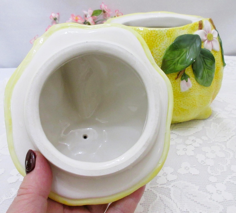May include: A ceramic lemon-shaped jar with a removable lid. The exterior features a textured yellow rind, green leaves, and small white blossoms. The interior of the lid and the jar are glazed in a smooth, glossy white finish with a small drainage hole.