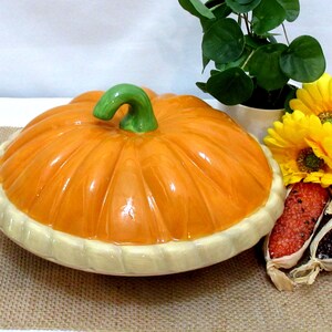 以下が含まれることがあります： オレンジ色のボディと緑色の茎が付いた、カボチャ型のセラミック製サービングディッシュ。お皿にはクリーム色の縁があります。黄色い花と小さな鉢植えが皿の隣にあります。お皿の直径は約30cmです。