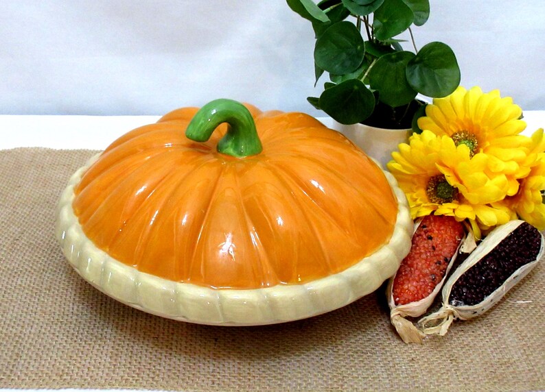 以下が含まれることがあります： オレンジ色のボディと緑色の茎が付いた、カボチャ型のセラミック製サービングディッシュ。お皿にはクリーム色の縁があります。黄色い花と小さな鉢植えが皿の隣にあります。お皿の直径は約30cmです。