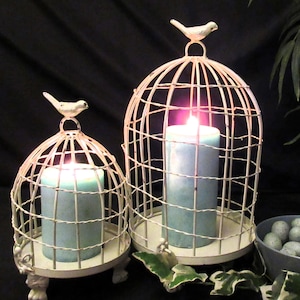 以下が含まれることがあります： 白い金属製の鳥かごが2つ、中にろうそくが入っています。鳥かごの上には鳥がいて、足つきの台の上に立っています。大きな鳥かごにはターコイズブルーのろうそく、小さな鳥かごには青いろうそくが入っています。