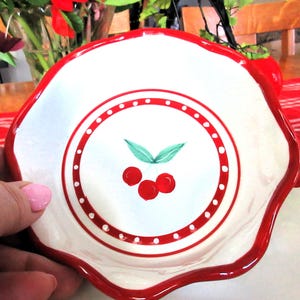 May include: A white ceramic dish with a scalloped edge and a red border. The centre features a cherry design with three red cherries and green leaves. A red dotted circle surrounds the cherry design. The dish is held by a person.