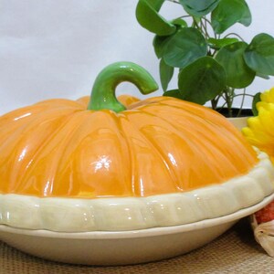 以下が含まれることがあります： 明るいオレンジ色の蓋と緑色の茎が付いた、カボチャ型のセラミック製サービングディッシュ。 ディッシュはクリーム色のベースとトリムです。 背景には、黄色い花、小さな植物、装飾用のトウモロコシが含まれています。