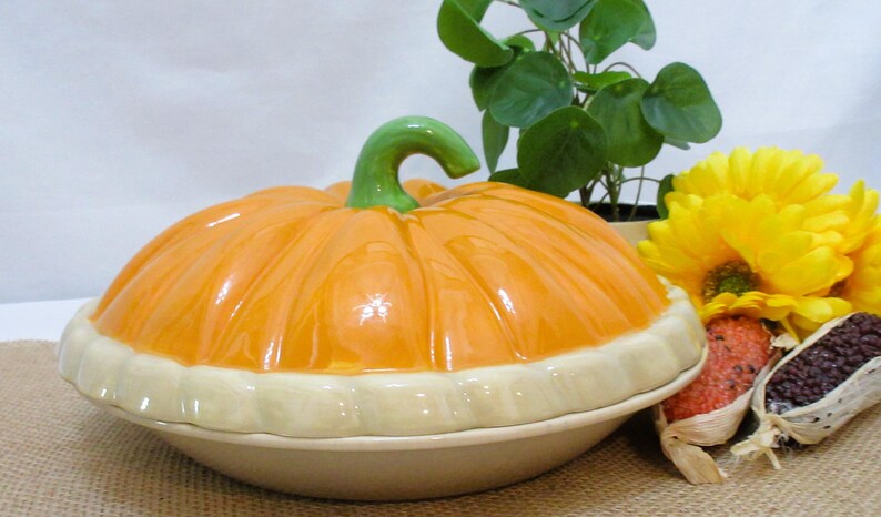 以下が含まれることがあります： 明るいオレンジ色の蓋と緑色の茎が付いた、カボチャ型のセラミック製サービングディッシュ。 ディッシュはクリーム色のベースとトリムです。 背景には、黄色い花、小さな植物、装飾用のトウモロコシが含まれています。