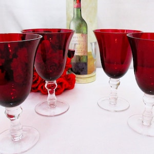 May include: Four ruby red wine glasses with clear stems and bases are arranged on a white surface. The glasses have a tulip shape. A bottle of wine and red roses are in the background.