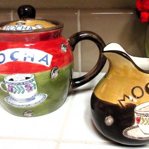 May include: A ceramic mocha teapot and creamer set. The teapot is red, green, and yellow with the word "MOCHA" and a coffee cup design. The creamer is brown and yellow with the word "MOCHA" and a coffee cup design.
